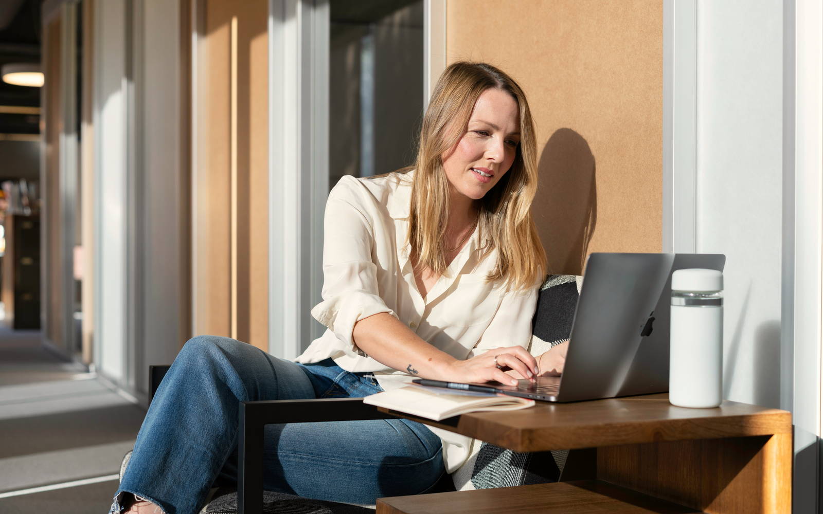Kvinna sitter vid ett träbord i solljus och arbetar vid datorn med ett anteckningsblock bredvid sig.
